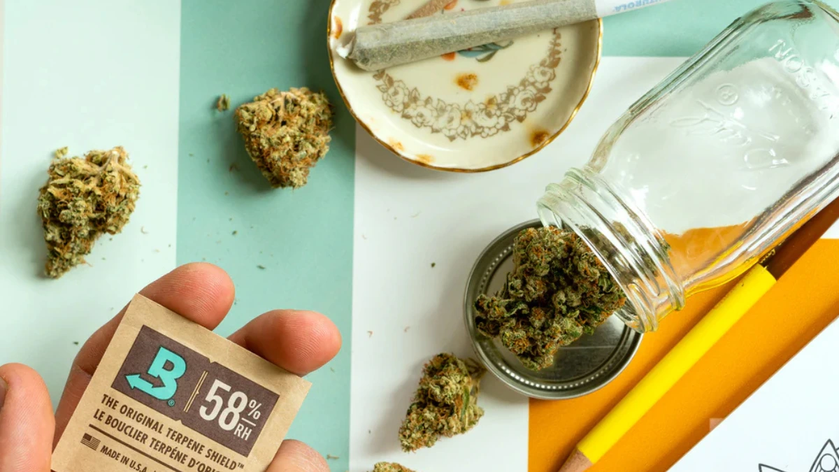 hand adding a two way humidity pack to a glass jar of cannabis with a pre-roll on the countertop