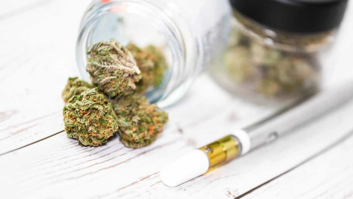 A cannabis vape pen next to a small jar of cannabis flower on a wooden surface.